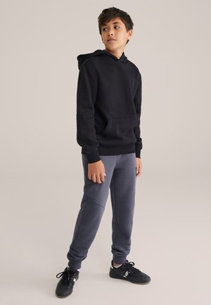 Schwarzer Kapuzenpullover mit einer Fronttasche und gerippten Bündchen, kombiniert mit grauen Jogginghosen mit Teilungsnähten, getragen mit schwarzen Sneakers.