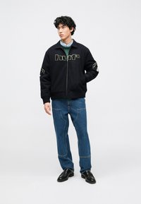 Jaqueta preta com fecho de correr, apresenta o logotipo "huf" em branco e cinzento, com bolsos laterais. Usada com jeans azuis e sapatos pretos.