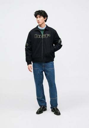 Giacca nera con zip caratterizzata dal logo "huf" in bianco e grigio, con tasche laterali. Indossata con jeans blu e scarpe nere.