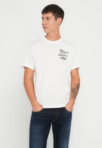 Camiseta de algodón blanca con un diseño gráfico negro que incluye el texto "Chrome and Leather", de mangas cortas y cuello redondo. Combinada con jeans de mezclilla oscuros.