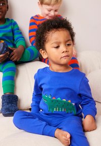 Blå pyjamas sæt med et grønt stegosaurus design på skjorten. Stoffet er blødt, og bukserne har en tætsiddende pasform. To børn i baggrunden.