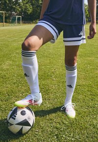 Adidas fotbollsskor i vitt med neonskott, i kombination med en svart-vit mönstrad fotboll, placerad på en gräsmatta.