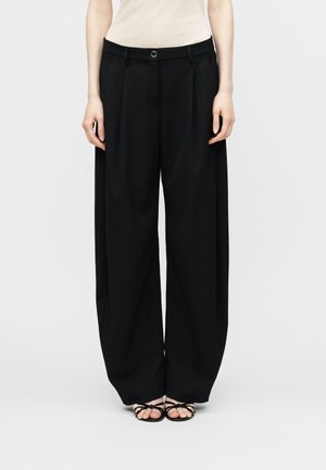 Kobieta w czarnych spodniach o szerokich nogawkach i czarnych sandałach na paski, stojąca na tle jednolitego jasnego tła.