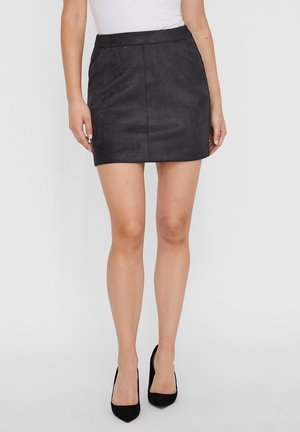 Mini jupe en faux daim noir avec une coupe droite, présentant une texture lisse et une taille ajustée. Associée à des escarpins noirs.