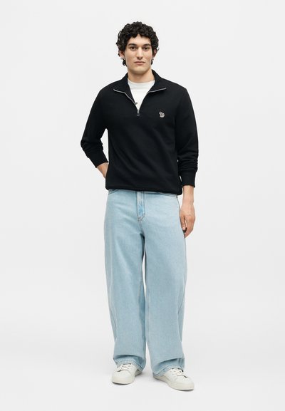 Νέος άνδρας που φοράει μαύρο πουλόβερ με φερμουάρ στο τέταρτο, ανοιχτό μπλε τζιν με φαρδιά παντελόνια και άσπρα αθλητικά παπούτσια, στέκεται μπροστά σε απλό λευκό φόντο.