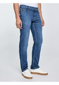 Vīrietis, kurš valkā zilas taisnas bikses un baltas sporta kurpes, stāv pret vienkrāsainu fonu.