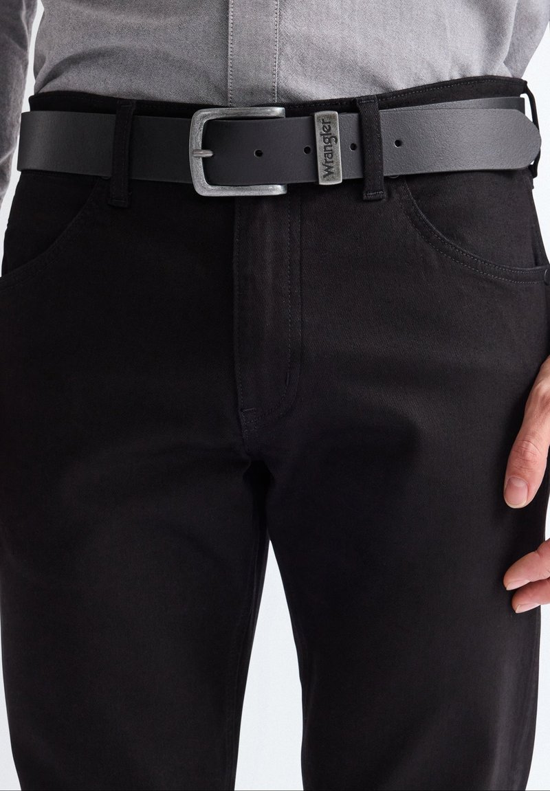 Man wearing black jeans and a black leather belt with a silver Wrangler buckle, with one hand resting near the pocket.