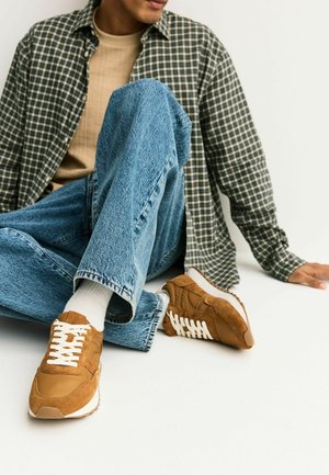 Person wearing blue jeans, beige sneakers with white laces, a beige shirt, and a green plaid shirt seated on the floor against a white background.