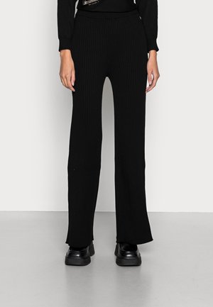 Frau trägt schwarze, gerippte, weit geschnittene Hosenbeine und schwarze Schuhe, steht vor einer schlichten hellen Wand und einem hellen Boden.