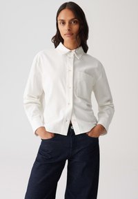 Vrouw met lang donker haar, gekleed in een wit overhemd met knopen en een borstzak, en donkerblauwe spijkerbroek, met handen in de zakken, tegen een effen achtergrond.