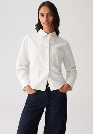 Vrouw met lang donker haar, gekleed in een wit overhemd met knopen en een borstzak, en donkerblauwe spijkerbroek, met handen in de zakken, tegen een effen achtergrond.