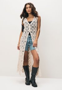 Pikk, lahtise eesosaga kardigan heledas beežis pitsis keerukate mustritega, kantud musta vardkatte ja teksapükste kohal, kombineeritud mustade põlvekõrguste saapadega.