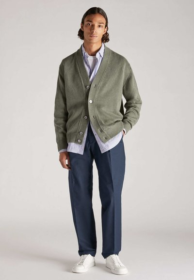 Un cárdigan verde de punto, abotonado y con bordes acanalados, usado sobre una camisa a rayas. Combinado con pantalones azul marino y zapatillas blancas.
