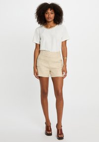 Hvit t-skjorte med korte ermer og perlebesetning ved halsen, kombinert med beige shorts med hundetannmønster. Brune hælsandaler fullfører antrekket.