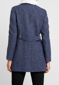 Kvinna som bär en blårandig ullkappa med krage av vit fårskinn och dekorativa knappar på baksidan av midjebandet.