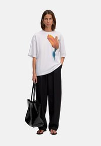 Weißes übergroßes T-Shirt mit orangefarbenem Tulpenmotiv, schwarze weite Hose und eine schwarze Tragetasche. Vor einem neutralen Hintergrund.