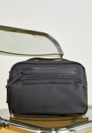 Sac bandoulière rectangulaire noir étanche avec deux compartiments zippés, placé sur une chaise transparente avec une structure métallique.