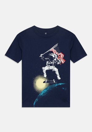 Chemise marine avec illustration d'un astronaute tenant un drapeau américain tout en faisant du skateboard au-dessus de la Terre avec le soleil en arrière-plan.