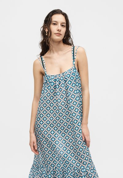 Γυναίκα με μακριά σκούρα μαλλιά που φοράει ένα αμάνικο φόρεμα σε μπλε, λευκό και καφέ γεωμετρικό σχέδιο με λεπτές τιράντες σε απλό φόντο.