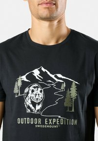 Sort bomulds t-shirt med et grafisk print af en bjørn, fyrretræer og bjerge i grøn og hvid. Teksten nedenunder lyder "OUTDOOR EXPEDITION SWIDEMOUNT."