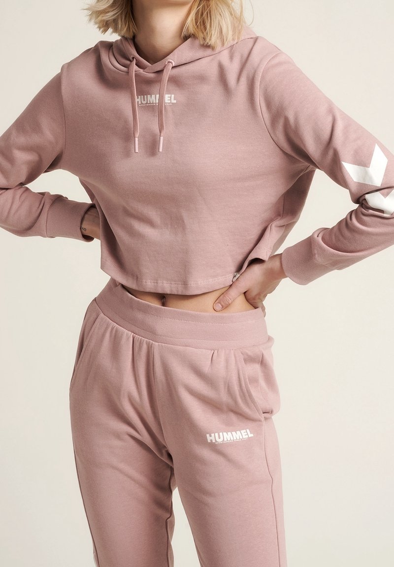 Femme portant un sweat à capuche court et un pantalon de jogging Hummel rose poudré assortis, mains sur les hanches, sur un fond neutre.