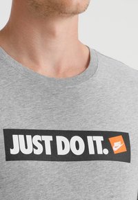 Grå bomullströja med ett brett svart rektangulärt grafiskt motiv med texten "JUST DO IT." och en orange Nike-logotyp i nedre högra hörnet.