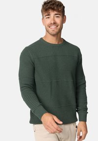 Pull en maille texturée vert foncé avec un col rond, présentant des motifs en côtes horizontales et en tissu gaufré, ainsi que des poignets ajustés.