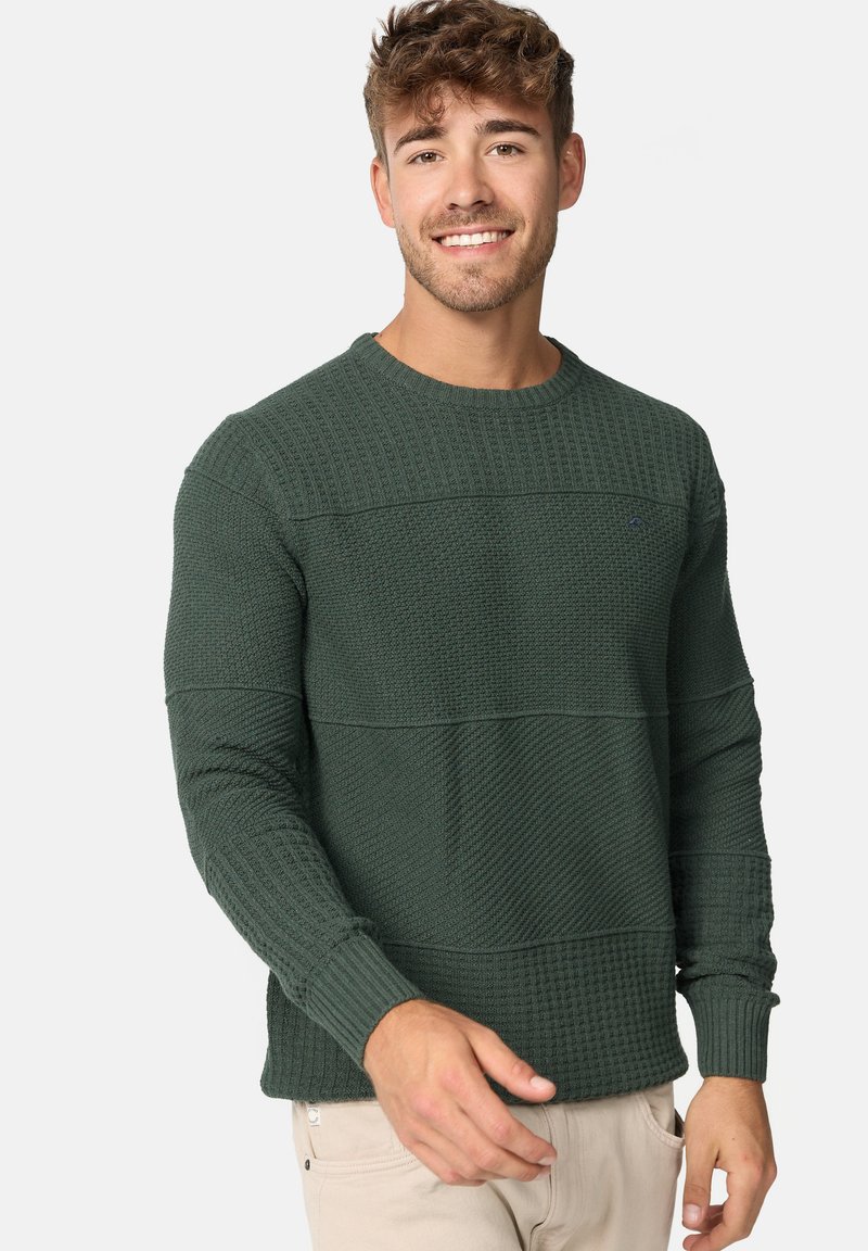 Pull en maille texturée vert foncé avec un col rond, présentant des motifs en côtes horizontales et en tissu gaufré, ainsi que des poignets ajustés.
