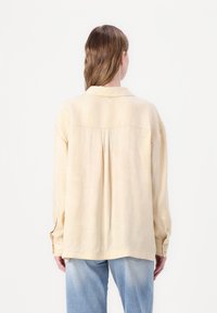 Beige, teksturert skjorte med krage, lange ermer og knappede mansjetter; har en bakfold og en rund knapp aksent nær halslinjen.