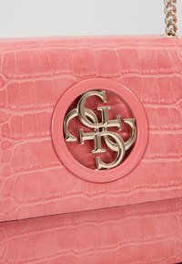 Różowa torebka o fakturze krokodyla z złotym, splecionym logo i łańcuszkiem na białym tle.