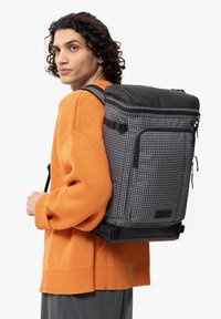 Zaino nero con motivo a griglia, caratterizzato da una forma strutturata, spallacci imbottiti e un lembo superiore con chiusura a zip.