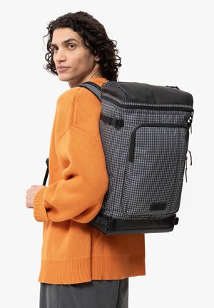 Backpack in black with a grid pattern, featuring a structured shape, padded shoulder straps, and a top flap with a zip closure.