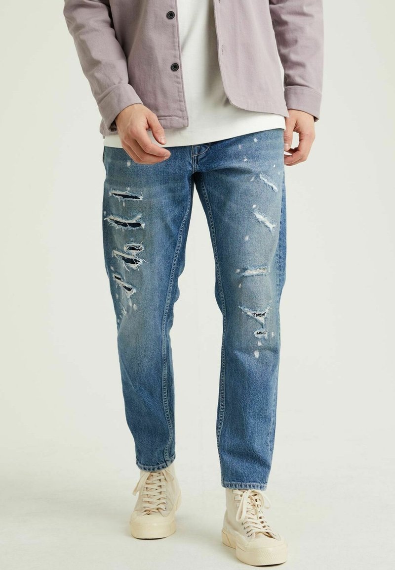 Jean en denim bleu à coupe décontractée, avec plusieurs patchs usés et ourlets effilochés, porté avec des baskets de couleur claire.