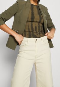 Zielono-oliwkowa blazerka o strukturalnym kroju z kieszenią na zamek, w połączeniu z ciemną, tie-dye, prążkowaną bluzką oraz wysokim stanem, kremowymi spodniami typu wide-leg.