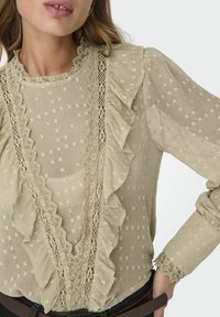 Heledbeez blouse, mille servade ja pitsdetailidega. Tekstuuriga kangas väikeste ruuduliste mustritega, pikad varrukad ja sügava V-kaelusega.