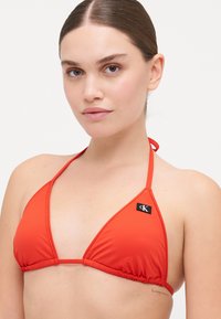 Röd triangulär bikiniöverdel i glatt tyg, med tunna band och en liten CK-logotag på vänster sida.
