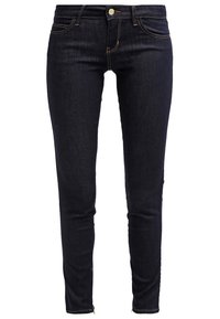 Vaqueros pitillo de mezclilla azul oscuro con una textura suave, diseño de cinco bolsillos y detalles de costuras doradas, con cremalleras en los tobillos.