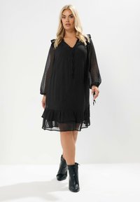 Schwarzes Mini-Kleid mit Punkten, V-Ausschnitt, Rüschendetails an den Ärmeln und gestuftem Saum. Kombiniert mit schwarzen Knöchelstiefeln.