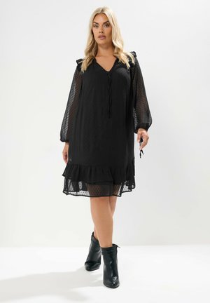 Schwarzes Mini-Kleid mit Punkten, V-Ausschnitt, Rüschendetails an den Ärmeln und gestuftem Saum. Kombiniert mit schwarzen Knöchelstiefeln.