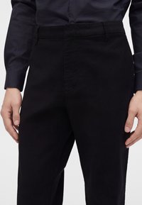 Personne portant un pantalon noir et une chemise sombre à manches longues, les mains relâchées le long du corps sur un fond clair.