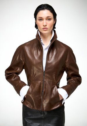 Mujer con cabello corto y oscuro, vestida con una chaqueta de cuero marrón con cremallera sobre una camisa blanca, con las manos en los bolsillos, frente a un fondo liso y claro.