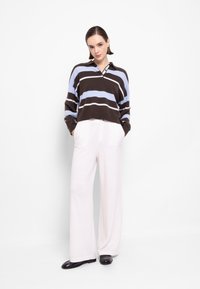 Brązowy i niebieski sweter w paski z kołnierzykiem, noszony z białymi spodniami o szerokich nogawkach i czarnymi butami. Gładka tekstura, luźny krój.