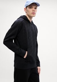 Μαύρη κουκούλα με υφασμάτινη υφή και τσέπη καγκουρό. Διαθέτει ρυθμιζόμενη κουκούλα με κορδόνια, ριμπ μανσέτες και στρίφωμα. Συνδυάζεται με μαύρο παντελόνι.