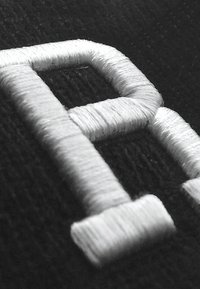 Hvit brodert bokstav "P" på en svart stoffbakgrunn, med teksturert søm og et litt hevet design.