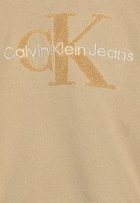 Felpa beige con ricami in oro testurizzati della scritta "CK" e "Calvin Klein Jeans" in bianco. Tessuto morbido con una finitura liscia.