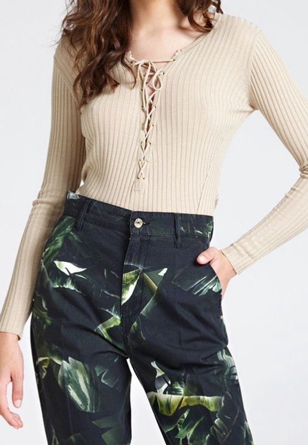 Femme portant un haut beige côtelé à manches longues avec un col à lacets et un pantalon noir taille haute imprimé de feuilles tropicales vertes.