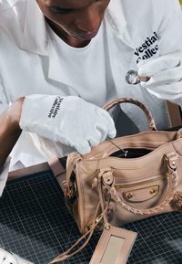 Personne portant des gants blancs inspecte le contenu d'un sac à main en cuir marron clair avec une petite loupe.
