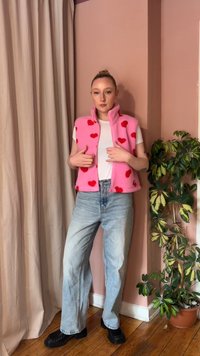 Mladá žena oblečená v ružovej flísovej veste s červenými srdiečkami, bielom tričku, svetlomodrých džínsoch a čiernych topánkach, stojaca pri ružovej stene s rastlinami.