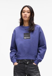Purple sweatshirt with a crew neck, ribbed cuffs, and hem. Features a black square logo patch with white text on the front.