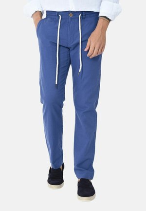 Uomo che indossa pantaloni casual blu con coulisse bianche, camicia bianca e scarpe slip-on nere, mani parzialmente nelle tasche.